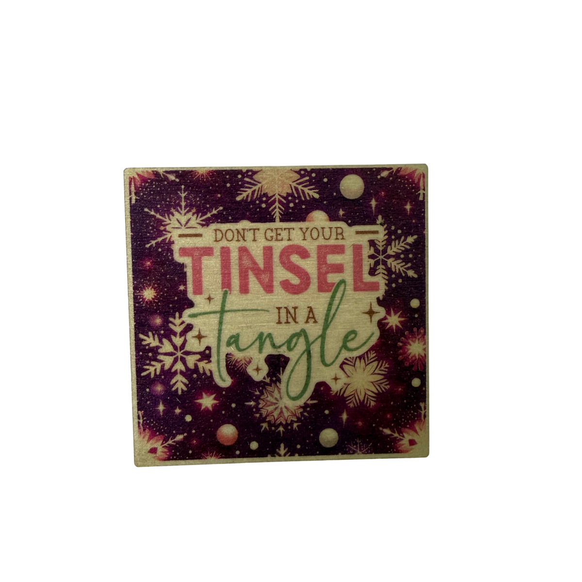 Square wooden Christmas magnet with Don't Get Your Tinsel in a Tangle lettering on a deep purple background with snowflakes and stars.