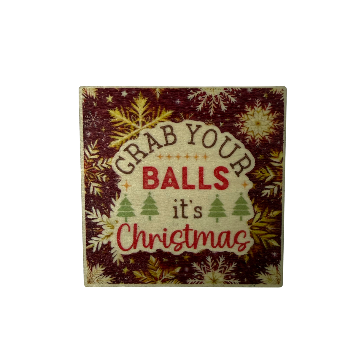 Square wooden Christmas magnet with Grab Your Balls It's Christmas lettering, green Christmas trees and snowflakes on a deep red background.